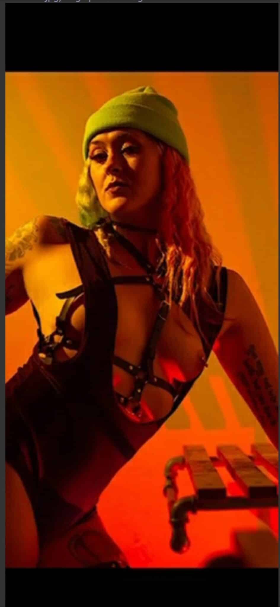 Bitch Flashing Pics: Studio Session: Unveiled Performance A woman performs on a synthesizer in a dimly lit studio, her bare shoulders and upper body exposed. Another figure watches from the background, creating an i...