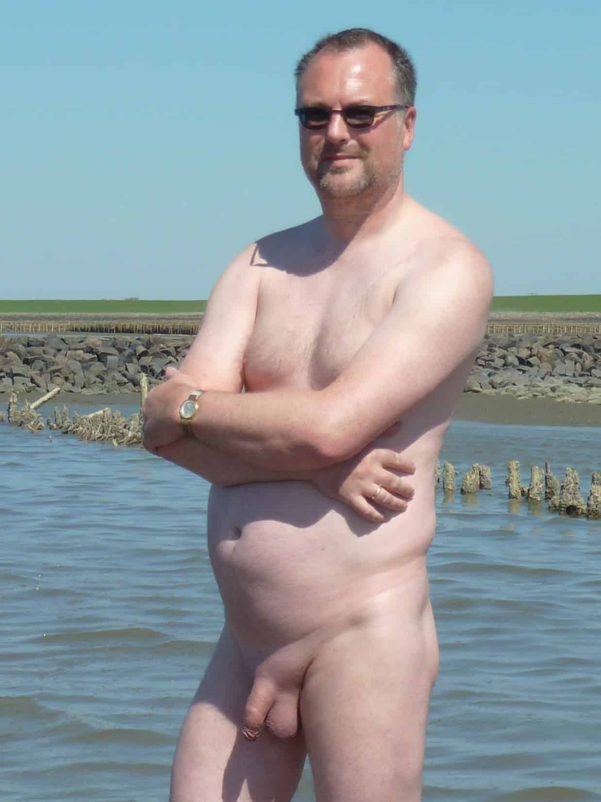 Dick Flash Pics: Why Is This Married Man Flashing His Cock at the Beach? A married man stands nude in shallow water, cock exposed, wearing only sunglasses and a wedding band, defying public nudity norms.